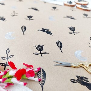May include: Brown paper with black ink stamps of flowers, leaves, and crescent moons. A pair of gold scissors is lying on the paper.