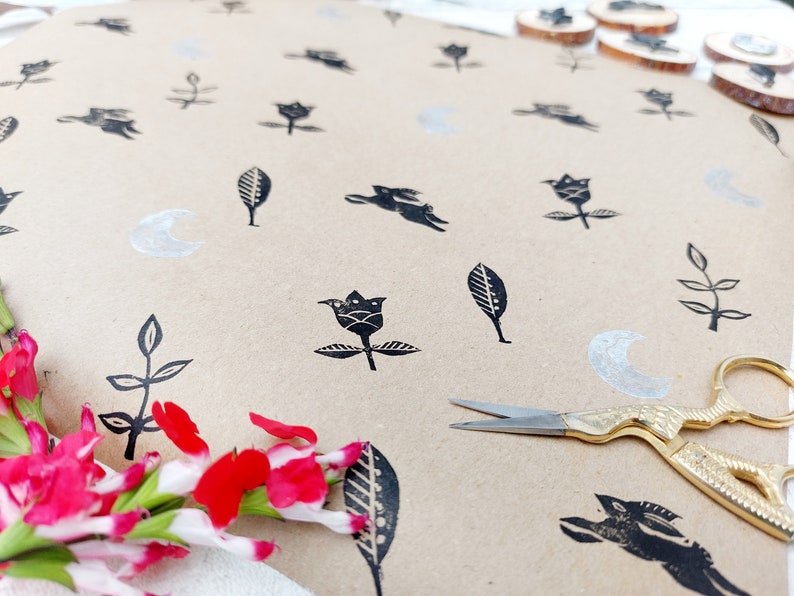 May include: Brown paper with black ink stamps of flowers, leaves, and crescent moons. A pair of gold scissors is lying on the paper.