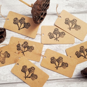 May include: Kraft paper gift tags with a black poppy seed head design. Each tag has a small hole for stringing. Several tags are scattered on a light wood-grain surface, along with dried seed pods.