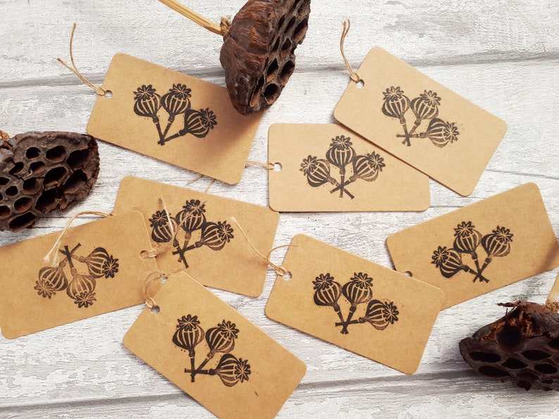 May include: Kraft paper gift tags with a black poppy seed head design. Each tag has a small hole for stringing. Several tags are scattered on a light wood-grain surface, along with dried seed pods.