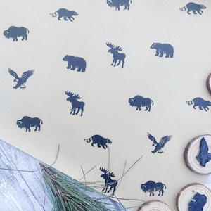 Puede incluir: Una hoja de papel de envolver marrón con un patrón repetitivo de siluetas negras de animales, incluyendo un oso, un bisonte, un alce, un águila y un mapache. El papel está sobre una superficie de madera con agujas de pino.