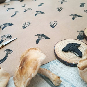 May include: A collection of mushroom-themed art supplies. The image features a variety of mushroom stamps in black ink on brown paper, along with real mushrooms and wooden slices. A pair of gold scissors and an ink pad are also visible.