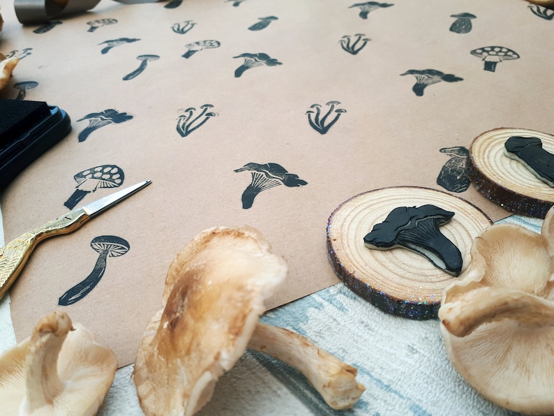 May include: A collection of mushroom-themed art supplies. The image features a variety of mushroom stamps in black ink on brown paper, along with real mushrooms and wooden slices. A pair of gold scissors and an ink pad are also visible.