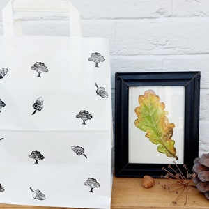 May include: White tote bag with black tree and acorn pattern. A framed leaf art piece with a black frame is next to the bag. A pinecone and acorn are also visible.