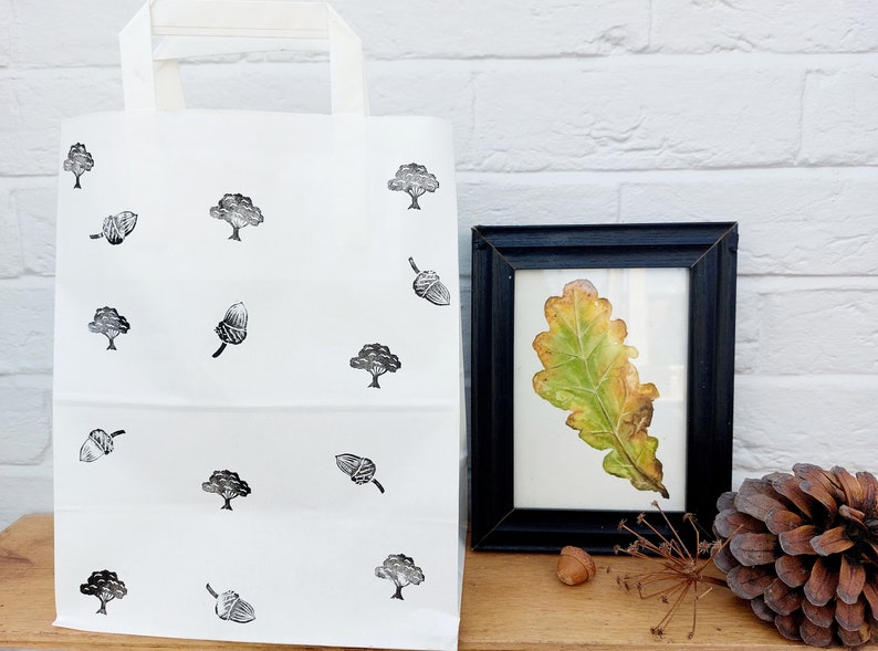 May include: White tote bag with black tree and acorn pattern. A framed leaf art piece with a black frame is next to the bag. A pinecone and acorn are also visible.
