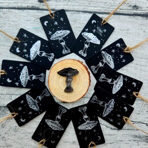 May include: A collection of black tags with white mushroom illustrations and star and moon accents, arranged around a wooden slice and a mushroom-shaped object. Each tag has a string attached for hanging.