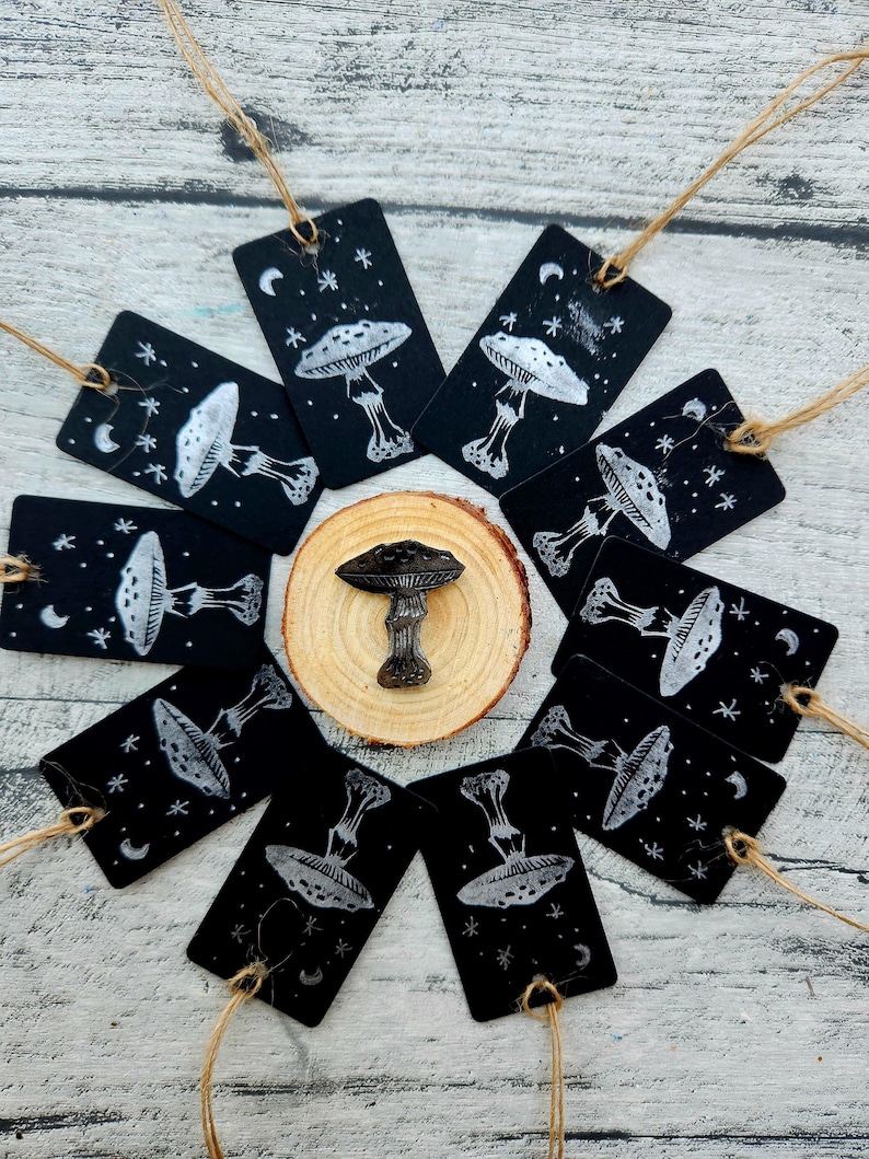 May include: A collection of black tags with white mushroom illustrations and star and moon accents, arranged around a wooden slice and a mushroom-shaped object. Each tag has a string attached for hanging.