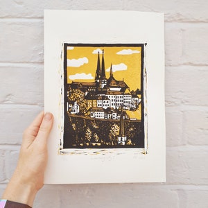 May include: A black and gold linocut print of a cityscape with a church in the center. The print is titled "Luxembourg City" and dated 2023.