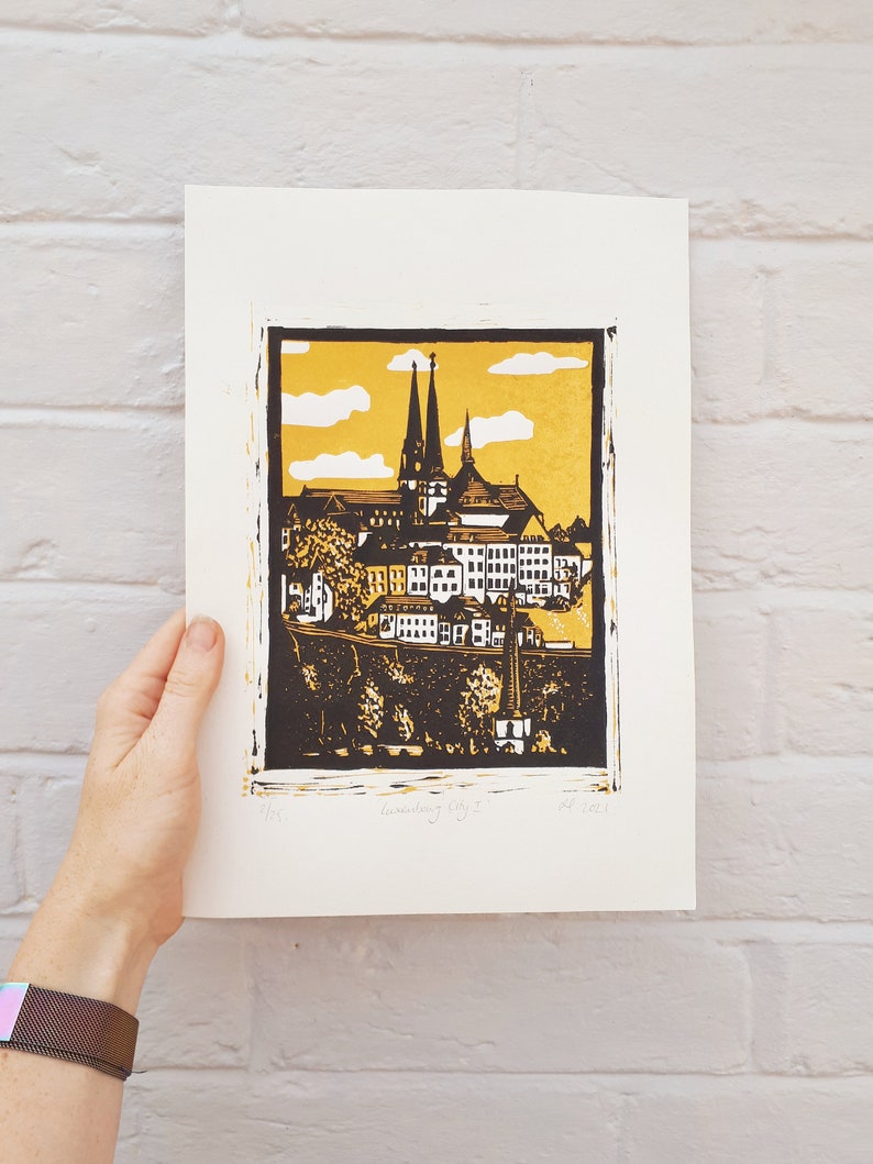 May include: A black and gold linocut print of a cityscape with a church in the center. The print is titled "Luxembourg City" and dated 2023.