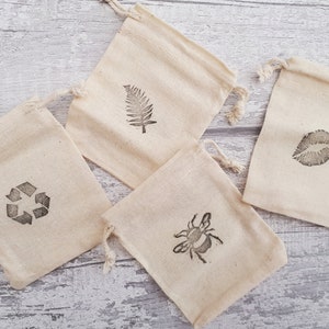 May include: Four small, white cotton drawstring bags with different designs printed on them. The designs include a fern leaf, a bee, a recycle symbol, and a pair of lips.