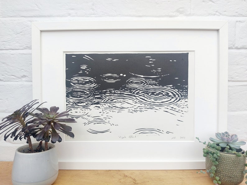May include: Black and white framed art print depicting the ripple effect of raindrops on water. The artwork is in a white frame and is displayed on a wooden surface. The print is next to a potted succulent.