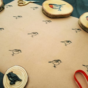 May include: A craft paper surface is covered with small bird illustrations. Wooden coasters with bird designs and a pair of gold scissors are also present. A red ribbon is visible in the lower right corner.