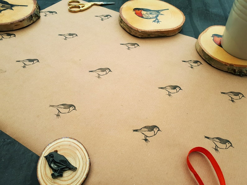May include: A craft paper surface is covered with small bird illustrations. Wooden coasters with bird designs and a pair of gold scissors are also present. A red ribbon is visible in the lower right corner.