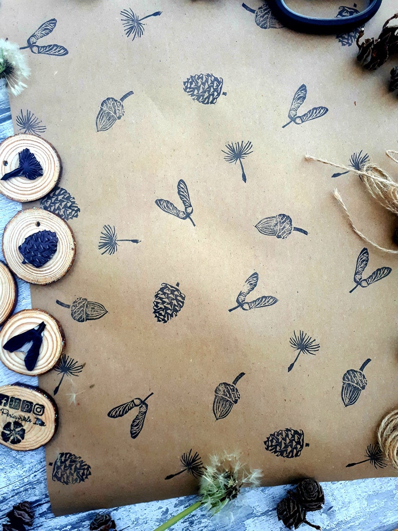 May include: Brown paper with a repeating pattern of black botanical illustrations, including acorns, pinecones, and seed pods. Wooden discs with similar stamped designs are arranged around the paper. A dandelion and twine are also visible.