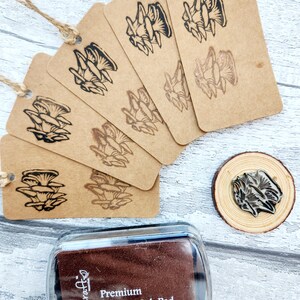 May include: A collection of kraft paper tags with a mushroom design stamped in black ink, strung with twine. A wooden stamp and a brown ink pad with the text "Premium Pigment Ink Pad" are also visible.