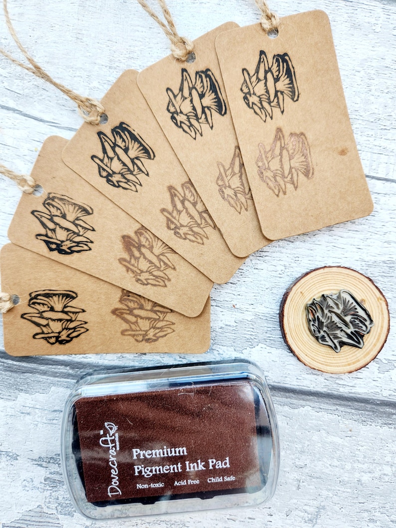 May include: A collection of kraft paper tags with a mushroom design stamped in black ink, strung with twine. A wooden stamp and a brown ink pad with the text "Premium Pigment Ink Pad" are also visible.