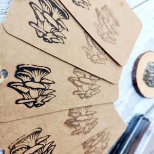 May include: A stack of brown paper tags with a black mushroom illustration. The tags are strung together with twine. The mushroom design is repeated on each tag. A small wooden slice is visible in the background.