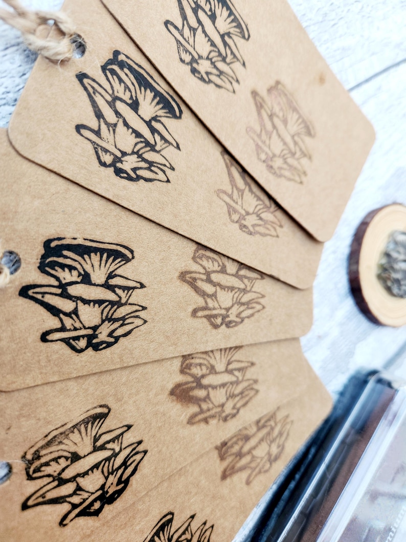 May include: A stack of brown paper tags with a black mushroom illustration. The tags are strung together with twine. The mushroom design is repeated on each tag. A small wooden slice is visible in the background.