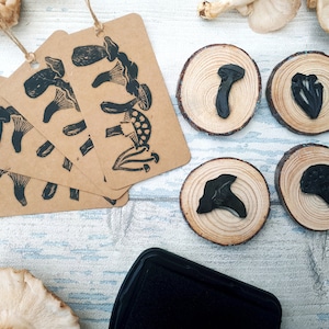 May include: A collection of craft supplies featuring mushroom-themed stamps and tags. Kraft paper tags with black mushroom illustrations hang with twine. Wooden rounds display stamped mushroom designs. A black ink pad sits nearby.