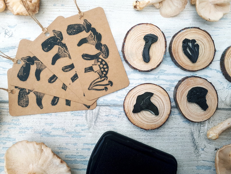 May include: A collection of craft supplies featuring mushroom-themed stamps and tags. Kraft paper tags with black mushroom illustrations hang with twine. Wooden rounds display stamped mushroom designs. A black ink pad sits nearby.