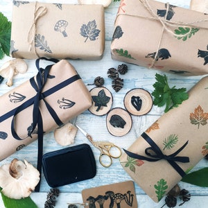 May include: Kraft paper-wrapped gifts with stamped botanical and woodland creature designs. The gifts are tied with twine and black ribbon. Additional elements include ink pads, scissors, and wooden slices with leaf and animal stamps.