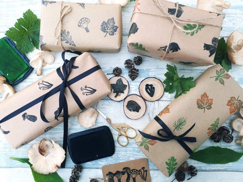 May include: Kraft paper-wrapped gifts with stamped botanical and woodland creature designs. The gifts are tied with twine and black ribbon. Additional elements include ink pads, scissors, and wooden slices with leaf and animal stamps.