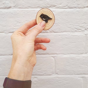 May include: A round wooden slice with a dark bird carving. The bird is perched on the wood, which has visible growth rings. The wood slice is held against a white brick wall. A dark metal mesh watch is on the wrist.