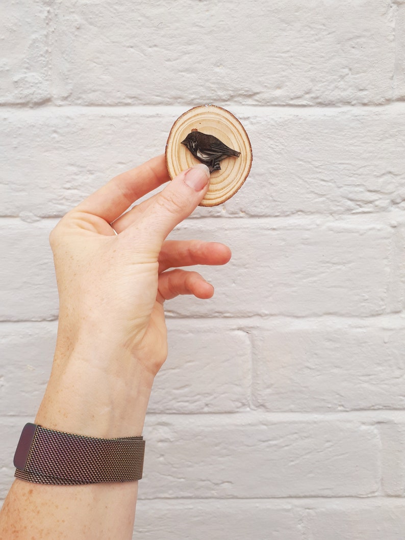May include: A round wooden slice with a dark bird carving. The bird is perched on the wood, which has visible growth rings. The wood slice is held against a white brick wall. A dark metal mesh watch is on the wrist.