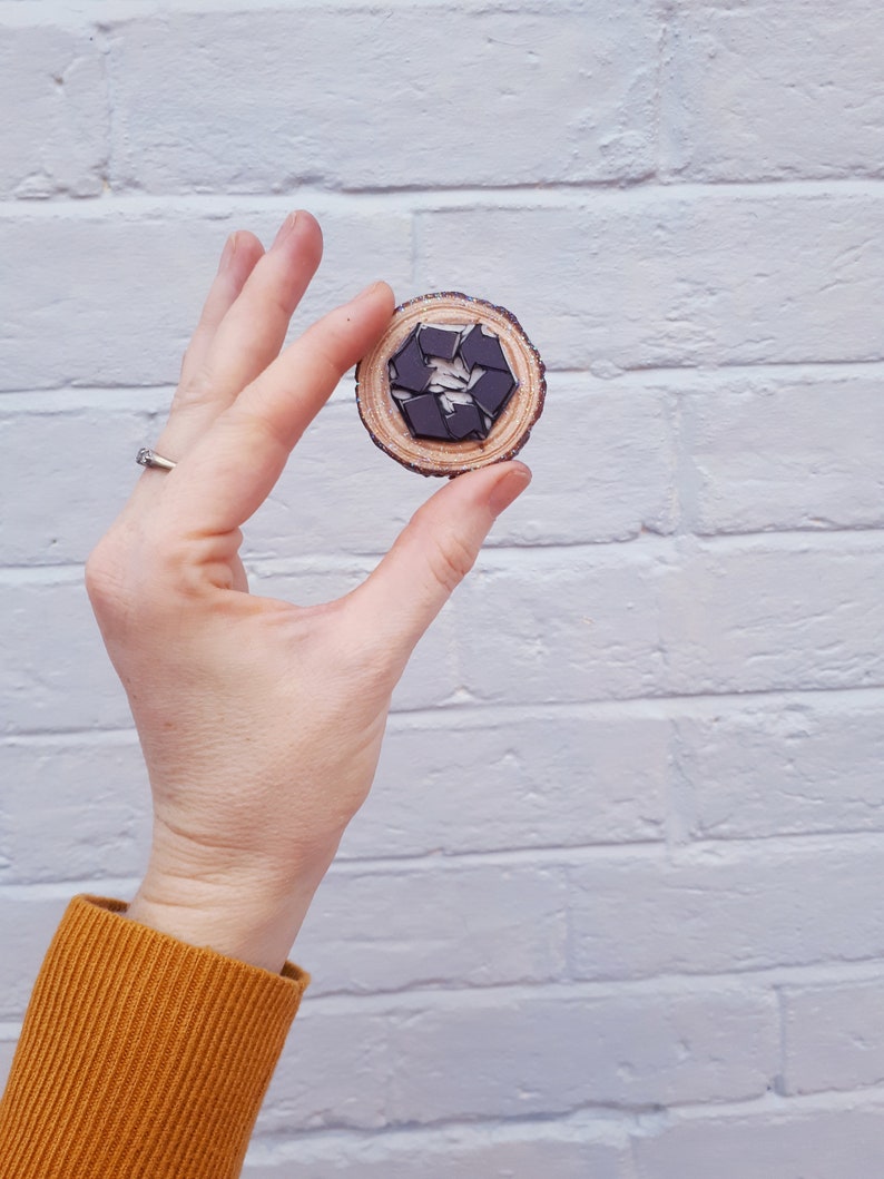 May include: A wooden circle with a black and white recycle symbol in the center. The circle is being held by a hand against a white brick wall.