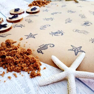 May include: A brown paper sheet with black ink stamps of various sea creatures, including starfish, seahorses, crabs, and jellyfish. There is a pile of brown sand and a white starfish on a white wooden surface.
