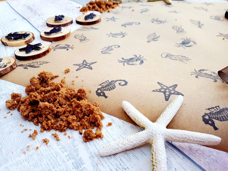 May include: A brown paper sheet with black ink stamps of various sea creatures, including starfish, seahorses, crabs, and jellyfish. There is a pile of brown sand and a white starfish on a white wooden surface.