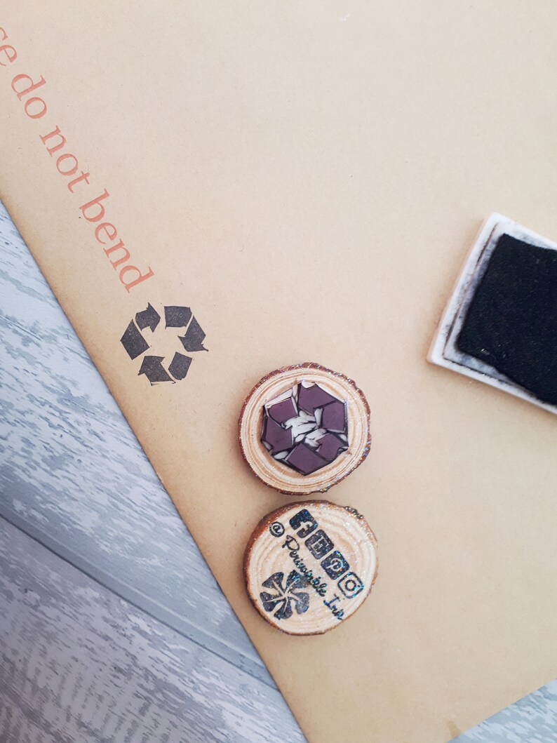 May include: Two wooden round stamps with a brown background. The first stamp has a black and white recycle symbol. The second stamp has a black and white logo with the text "AMPO" and "@Perishable Ink".
