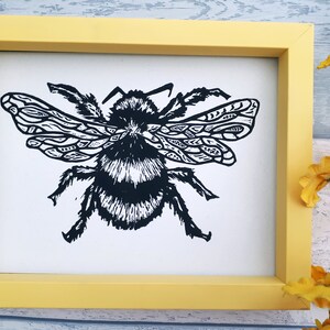 Puede incluir: Una impresi&oacute;n enmarcada que presenta una ilustraci&oacute;n en lin&oacute;leo en blanco y negro de un abejorro con patrones geom&eacute;tricos intrincados en sus alas. La obra est&aacute; encerrada en un marco rectangular amarillo s&oacute;lido, presentando un dise&ntilde;o gr&aacute;fico de alto contraste.