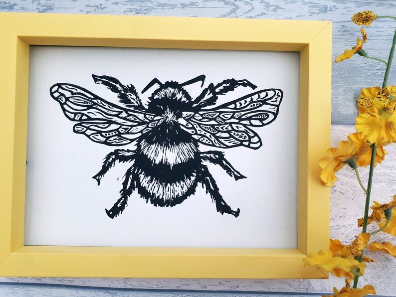 Puede incluir: Una impresi&oacute;n enmarcada que presenta una ilustraci&oacute;n en lin&oacute;leo en blanco y negro de un abejorro con patrones geom&eacute;tricos intrincados en sus alas. La obra est&aacute; encerrada en un marco rectangular amarillo s&oacute;lido, presentando un dise&ntilde;o gr&aacute;fico de alto contraste.