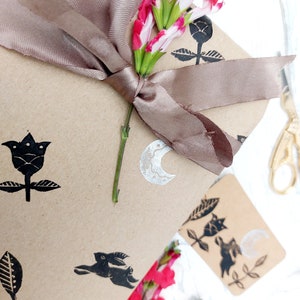 May include: A brown paper gift bag with a brown satin ribbon tied around it. The bag is decorated with black and white stamps of flowers, leaves, a crescent moon, and a rabbit. A bouquet of pink and white flowers is on top of the bag.