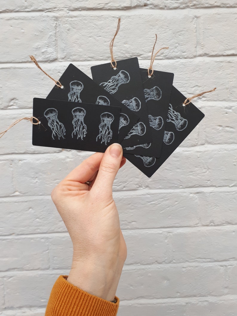 May include: A set of six black gift tags with white ink illustrations of jellyfish. The tags are tied with twine.
