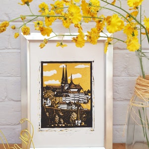 May include: A framed black and white linocut print of a cityscape with a gold background. The print depicts a city with tall buildings and a church steeple. The print is titled "Landsberg City" and is signed and dated.
