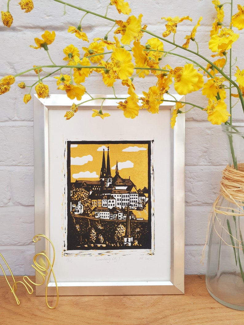 May include: A framed black and white linocut print of a cityscape with a gold background. The print depicts a city with tall buildings and a church steeple. The print is titled "Landsberg City" and is signed and dated.