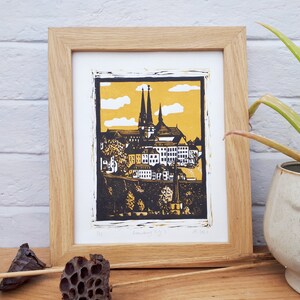 May include: A framed black and yellow linocut print of a cityscape with a church in the background. The print is titled "Linocut City" and dated "May 2021".