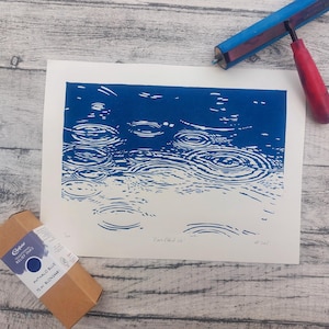 May include: A blue and white print depicting raindrops on water. A red and blue printmaking brayer and a box of blue relief ink are also present. The print is on a white background, and the ink box is brown.