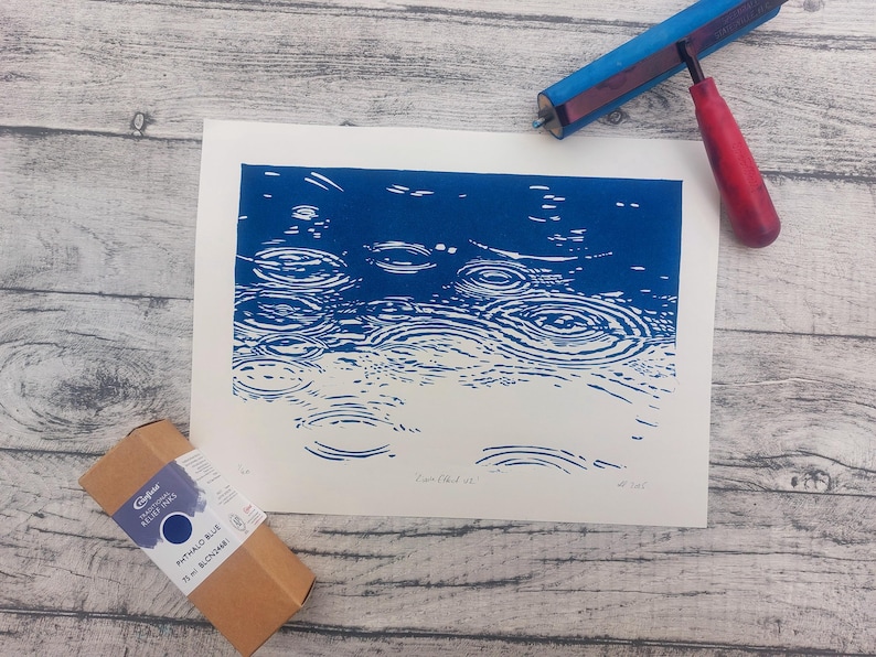 May include: A blue and white print depicting raindrops on water. A red and blue printmaking brayer and a box of blue relief ink are also present. The print is on a white background, and the ink box is brown.