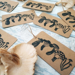 May include: Kraft paper tags with a black mushroom linocut design. The tags are rectangular with a string for hanging. Several tags are arranged with dried mushrooms on a light blue wooden surface.