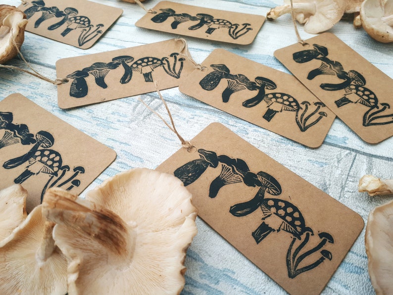 May include: Kraft paper tags with a black mushroom linocut design. The tags are rectangular with a string for hanging. Several tags are arranged with dried mushrooms on a light blue wooden surface.