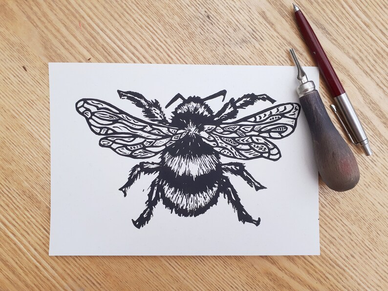 Puede incluir: Una impresi&oacute;n en tinta negra de un abejorro sobre papel rectangular blanco. La abeja presenta alas con patrones detallados y un cuerpo peludo. Junto al papel hay una herramienta de grabado en lin&oacute;leo con mango de madera y un bol&iacute;grafo fino.