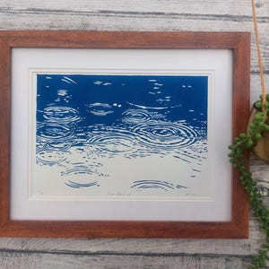 May include: Framed art print depicting a blue and white linocut of raindrops creating ripples on a water surface. The artwork is in a brown wooden frame with a white mat. A hanging plant in a gold pot is visible.