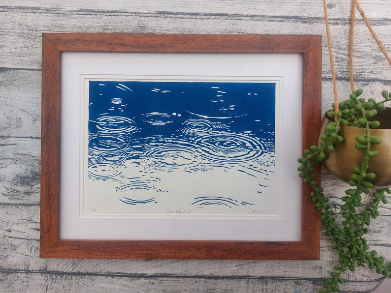 May include: Framed art print depicting a blue and white linocut of raindrops creating ripples on a water surface. The artwork is in a brown wooden frame with a white mat. A hanging plant in a gold pot is visible.