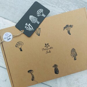 May include: A brown cardboard box with black mushroom illustrations and a black tag with white mushroom designs. The box has a round sticker with the text "Periwinkle Ink". The tag is attached with twine.