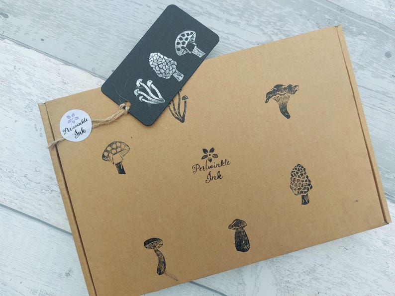 May include: A brown cardboard box with black mushroom illustrations and a black tag with white mushroom designs. The box has a round sticker with the text "Periwinkle Ink". The tag is attached with twine.