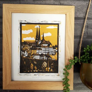 May include: A framed black and white linocut print of a cityscape with a church in the background. The print is on a yellow background with white clouds. The print is titled "Linocut City 1" and is dated 2021.