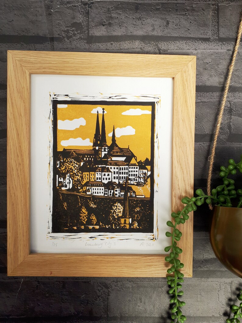 May include: A framed black and white linocut print of a cityscape with a church in the background. The print is on a yellow background with white clouds. The print is titled "Linocut City 1" and is dated 2021.